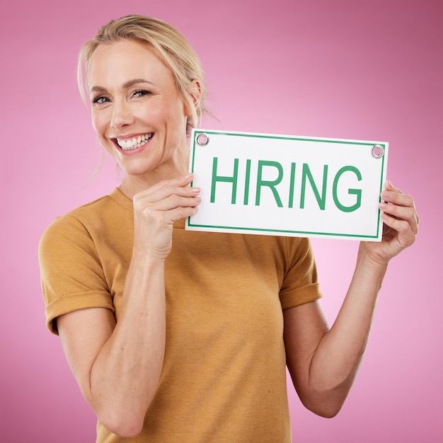 Hiring sign and portrait of a woman for recruitment job search and advertising opportunity Smile of a happy business person with a poster for recruiting and human resources on a pink background