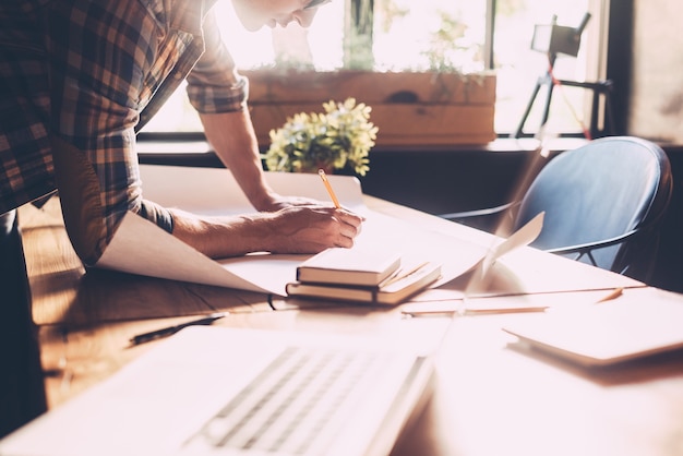 His job is his life. Close-up of confident young man in casual wear sketching on blueprint while standing near wooden desk in creative office