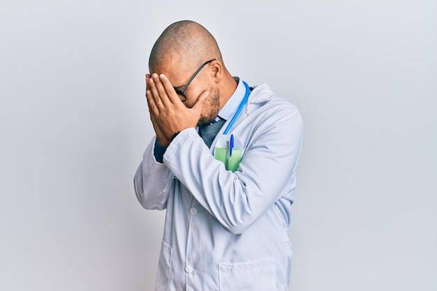 Hispanic adult man wearing doctor uniform and stethoscope with sad expression covering face with hands while crying depression concept