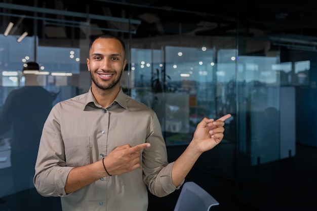 Hispanic businessman inside the office smiling and looking at the camera while standing and pointing