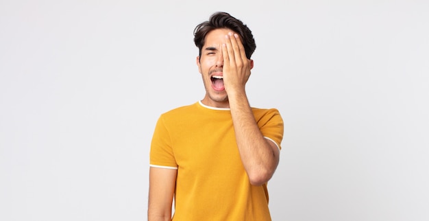 Hispanic handsome man looking sleepy, bored and yawning, with a headache and one hand covering half the face