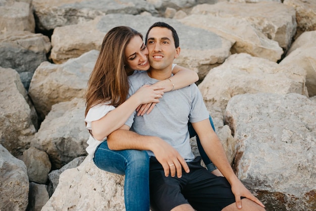 A Hispanic man is sitting with his girlfriend on the stones of a breakwater