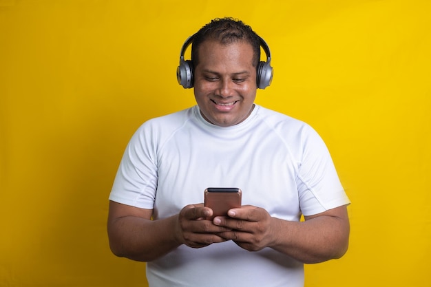 Hispanic man listening to music with headphones and cell phone, on yellow background