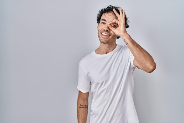 Hispanic man standing over isolated background doing ok gesture with hand smiling eye looking through fingers with happy face