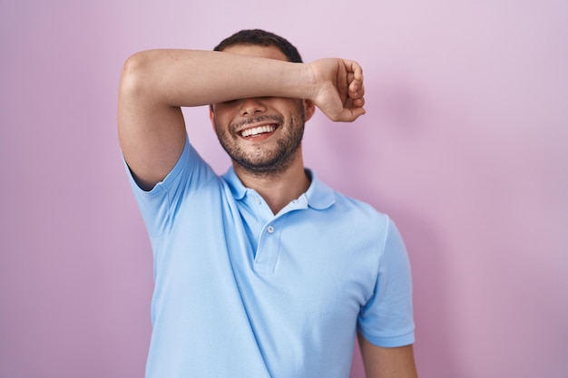 Hispanic man standing over pink background covering eyes with arm smiling cheerful and funny. blind concept.