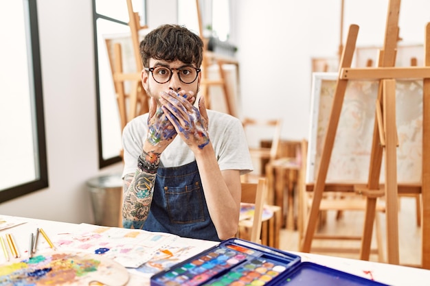 Hispanic man with beard at art studio shocked covering mouth with hands for mistake secret concept