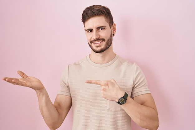 Hispanic man with beard standing over pink background amazed and smiling to the camera while presenting with hand and pointing with finger