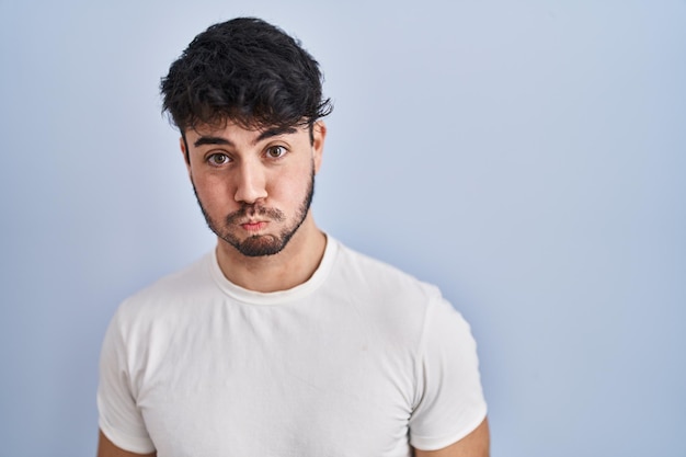 Hispanic man with beard standing over white background puffing cheeks with funny face. mouth inflated with air, crazy expression.