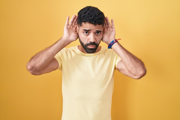 Photo hispanic man with beard standing over yellow background trying to hear both hands on ear gesture, curious for gossip. hearing problem, deaf