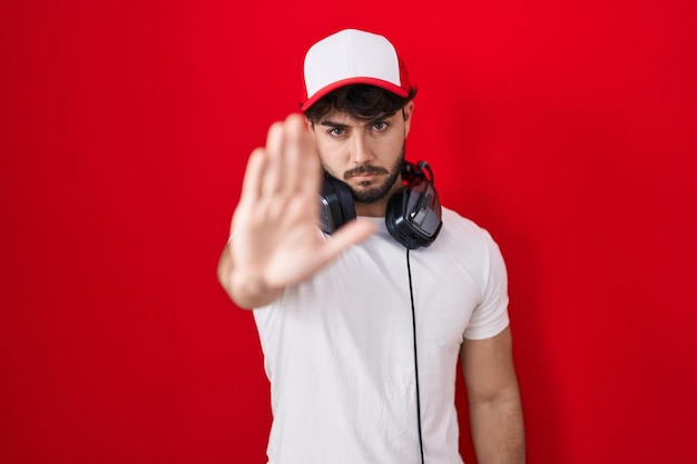 Hispanic man with beard wearing gamer hat and headphones doing stop sing with palm of the hand. warning expression with negative and serious gesture on the face.