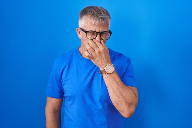 Hispanic man with grey hair standing over blue background smelling something stinky and disgusting intolerable smell holding breath with fingers on nose bad smell