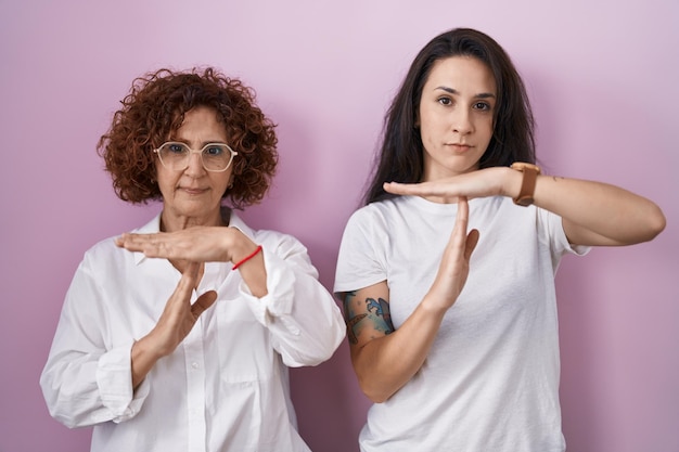 Hispanic mother and daughter wearing casual white t shirt over pink background doing time out gesture with hands, frustrated and serious face