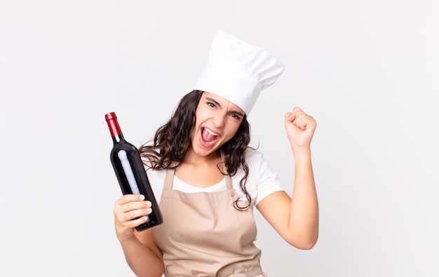 Hispanic pretty chef woman shouting aggressively with an angry expression and holding a wine bottle