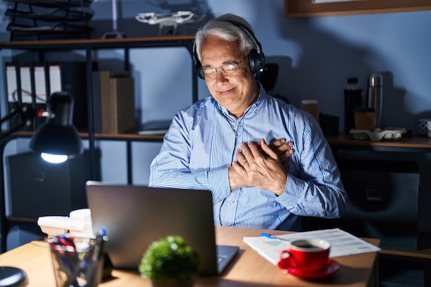 Hispanic senior man wearing call center agent headset at night smiling with hands on chest with closed eyes and grateful gesture on face. health concept.