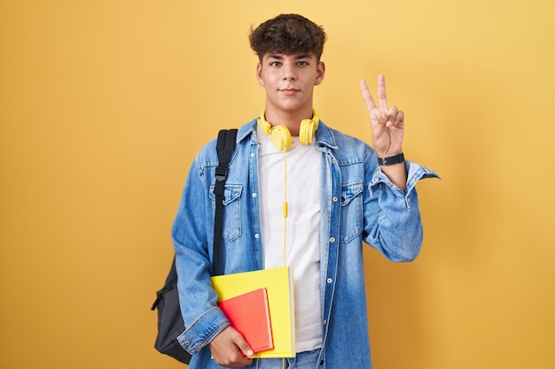 Hispanic teenager wearing student backpack and holding books smiling looking to the camera showing fingers doing victory sign number two