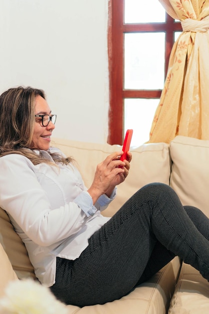 Hispanic woman lying on the couch at home She is holding a smartphone which she looks at and smiles