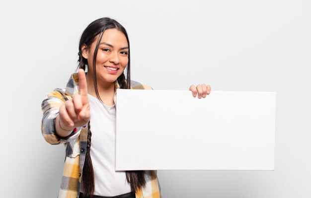 Hispanic woman smiling proudly and confidently making number one pose triumphantly, feeling like a leader