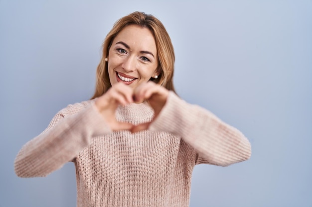 Hispanic woman standing over blue background smiling in love doing heart symbol shape with hands romantic concept