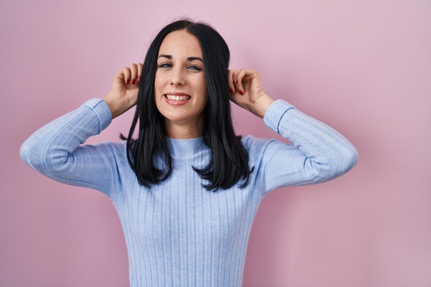 Hispanic woman standing over pink background smiling pulling ears with fingers funny gesture audition problem