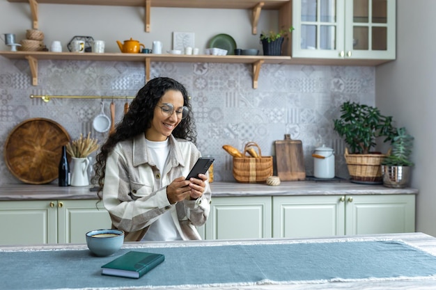 Hispanic woman uses mobile phone at home in the kitchen woman smiles and rejoices reads messages