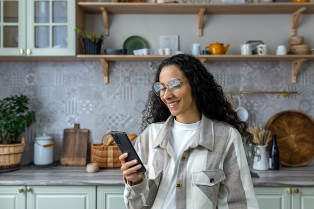 Hispanic woman uses mobile phone at home in the kitchen woman smiles and rejoices reads messages