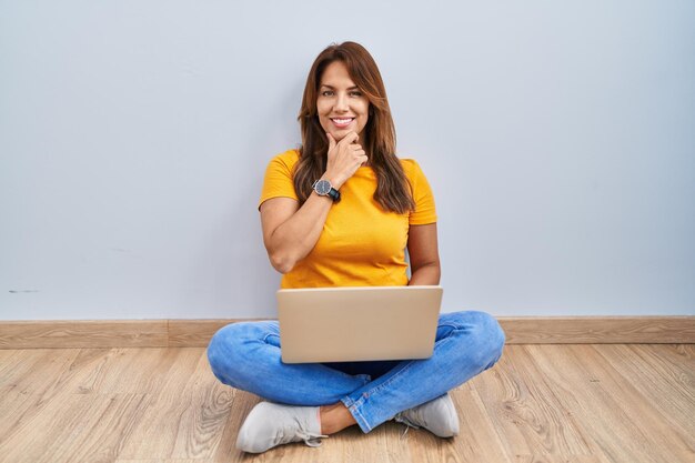 Hispanic woman using laptop sitting on the floor at home with hand on chin thinking about question, pensive expression. smiling and thoughtful face. doubt concept.