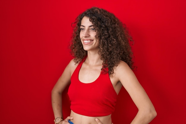 Hispanic woman with curly hair standing over red background looking away to side with smile on face, natural expression. laughing confident.