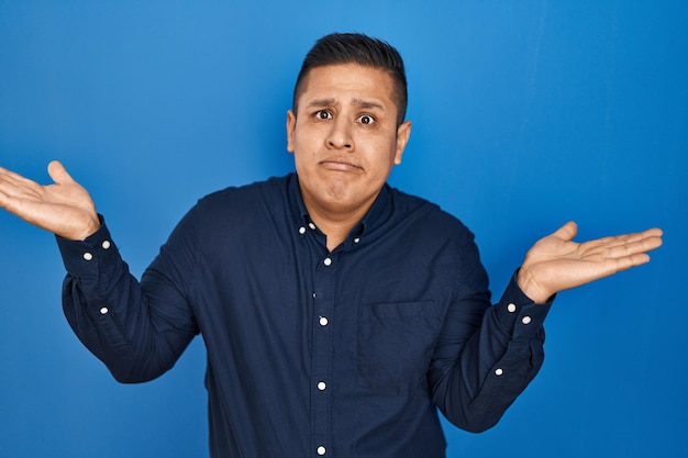 Hispanic young man standing over blue background clueless and confused expression with arms and hands raised. doubt concept.