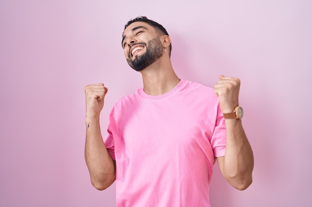 Hispanic young man standing over pink background celebrating surprised and amazed for success with arms raised and eyes closed. winner concept.