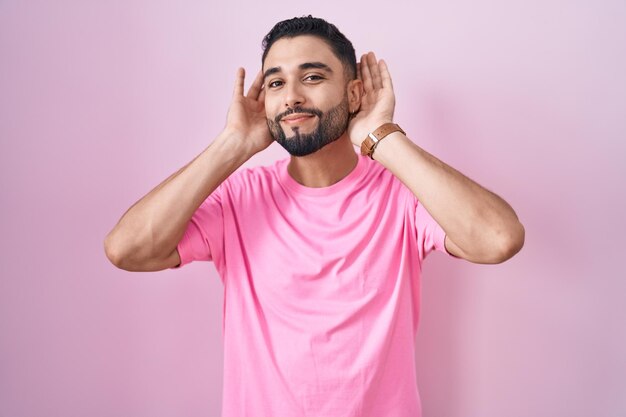 Photo hispanic young man standing over pink background trying to hear both hands on ear gesture curious for gossip hearing problem deaf