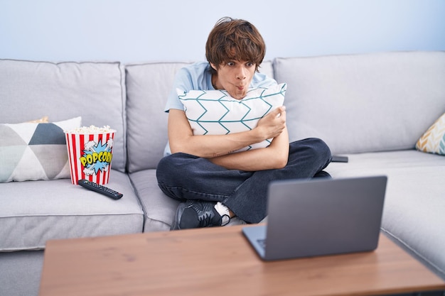 Hispanic young man watching a horror movie in the laptop making fish face with mouth and squinting eyes, crazy and comical.