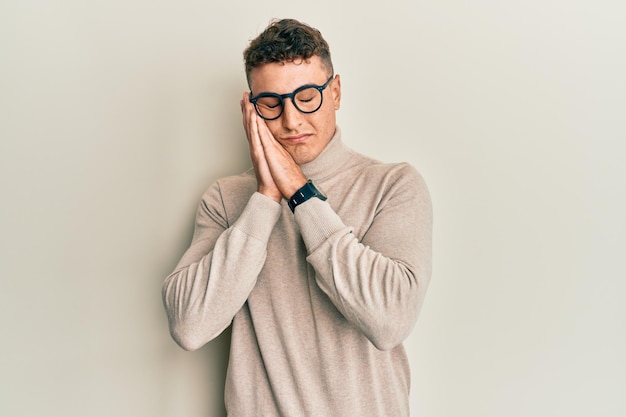 Hispanic young man wearing casual turtleneck sweater sleeping tired dreaming and posing with hands together while smiling with closed eyes