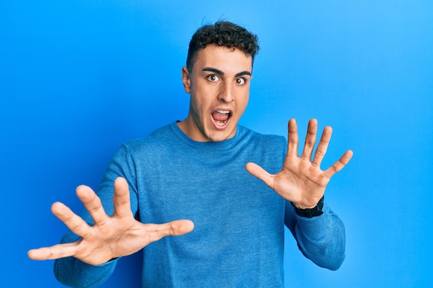 Hispanic young man wearing casual winter sweater afraid and terrified with fear expression stop gesture with hands shouting in shock panic concept