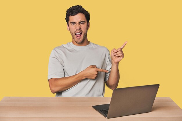 Photo hispanic young man working on laptop pointing with forefingers to a copy space