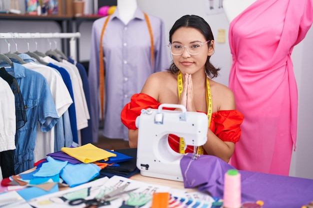 Hispanic young woman dressmaker designer using sewing machine praying with hands together asking for forgiveness smiling confident.