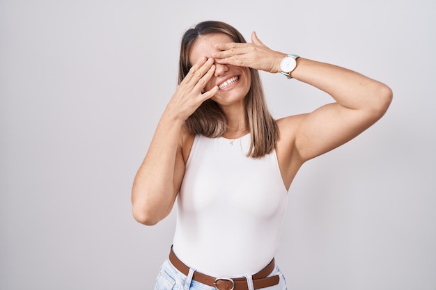 Hispanic young woman standing over white background covering eyes with hands smiling cheerful and funny. blind concept.