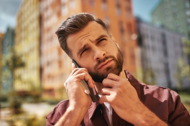 Hm... Portrait of thoughtful bearded man talking by phone and touching his stubble while standing on the street. Bad news. Digital. Communication