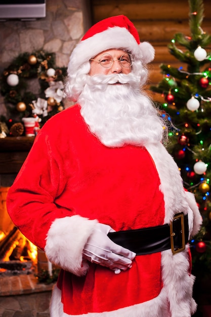 Ho Ho Ho! Cheerful Santa Claus adjusting his belt and smiling with fireplace and Christmas Tree in the background