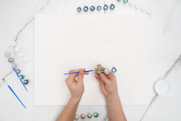 Hobby at home painting by numbers to draw from above a view of women's hands holding cans of paints.