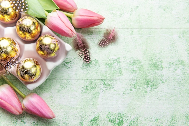 Holder with Easter eggs painted golden colors and pink tulips on a green wooden background