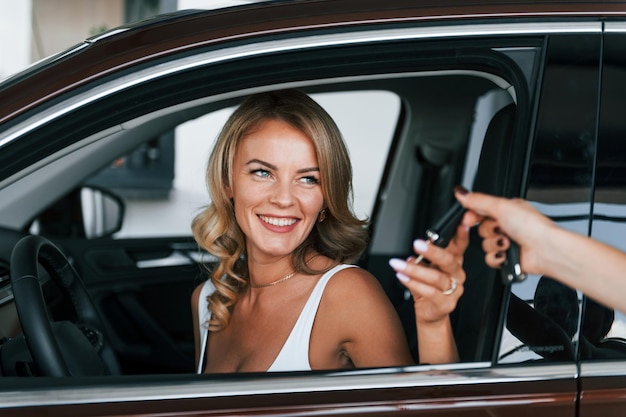 Holding car keys Woman in formal clothes is indoors in the autosalon