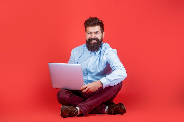 Holding laptop computer Smiling handsome bearded man worker laptop Happy young man sitting on the floor with and using laptop computer on red background