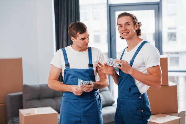 Holding money Two young movers in blue uniform working indoors in the room