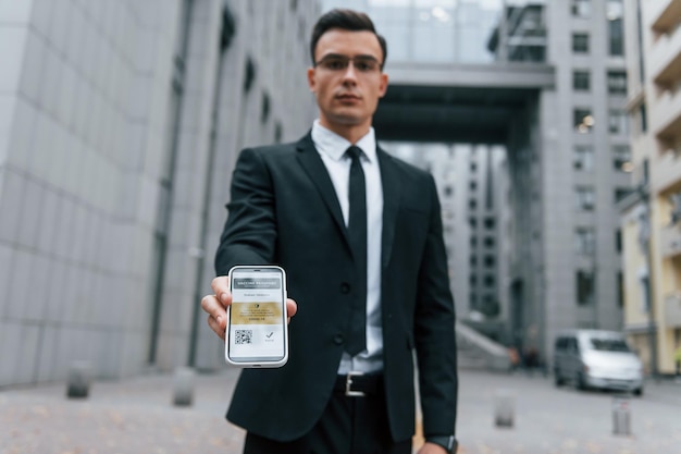 Holding phone with vaccination certificate Businessman in black suit and tie is outdoors in the city