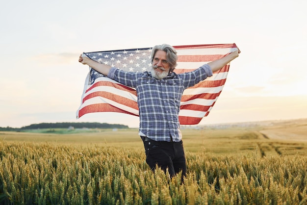 Holding USA flag in hands Patriotic senior stylish man with grey hair and beard on the agricultural field