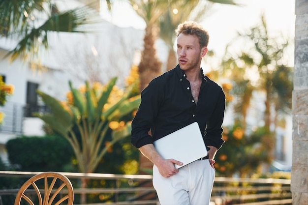 Holds laptop Young man is outdoors at sunny daytime Concept of vacation