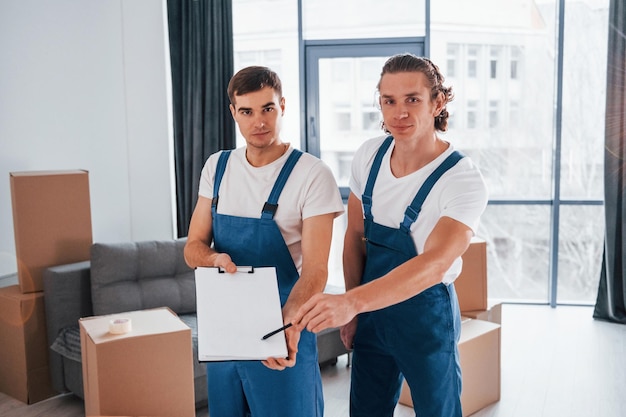 Holds notepad with document Two young movers in blue uniform working indoors in the room