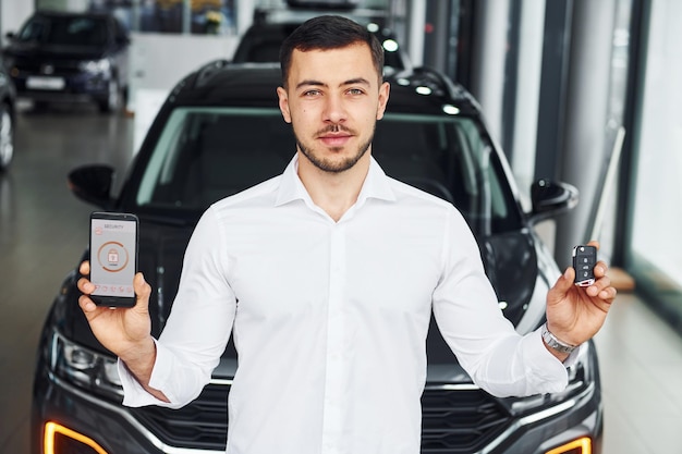 Holds phone with labels and icons Young man in white shirt is indoors with modern new automobile Remote control