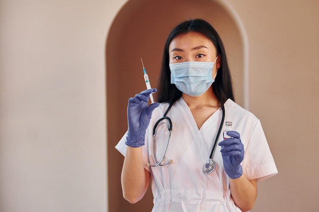 Holds vaccine and syringe Young serious asian woman standing indoors