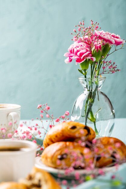 Holiday break with cup of coffee, mini fresh croissants chocolate bun and carnation flowers on turquoise surface. Copy space